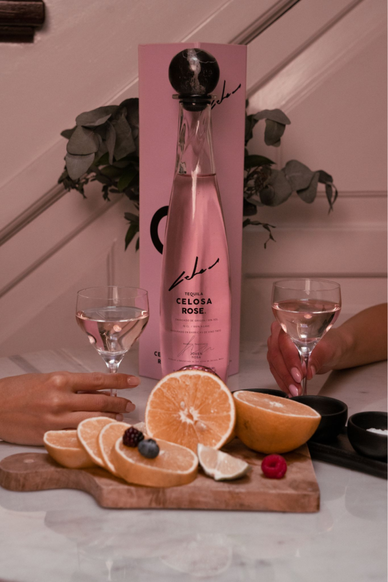 A bottle and two glasses of Celosa Rose Pink Tequila, and citrus fruits.