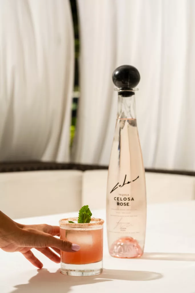 A hand reaching for a glass of Rose Margarita on a table, garnished with a sprig of mint and rimmed with pink salt. In the background, there is a bottle labeled "TEQUILA CELOSA ROSE" with a sleek design and black cap, partially obscured by a sheer white curtain.