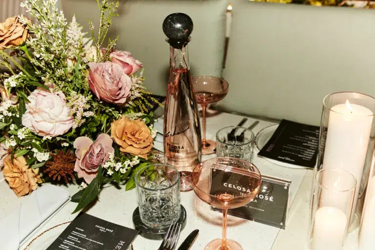 A table elegantly set with a bouquet of flowers in shades of pink and cream, a bottle of pink tequila labeled "CELOSA ROSE," and matching glasses half-filled with the same pink liquid. The table also features crystal clear drinking glasses, lit candles providing a warm ambiance, and menus with black print, creating a sophisticated and inviting presentation.