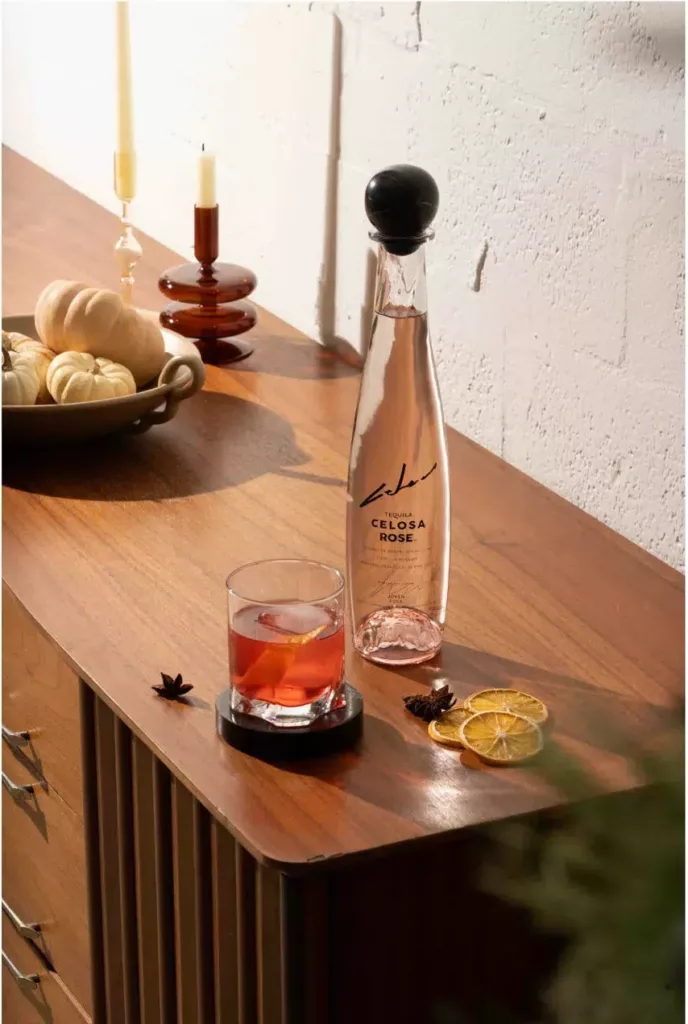 Celosa Rose Tequila bottle next to a Negroni cocktail with lemon and star anise on a wooden sideboard with elegant candles in the background.