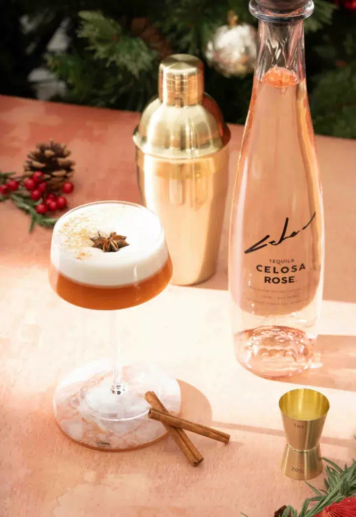 Stylish presentation of Celosa Rose Tequila bottle next to a frothy Spiced Sour cocktail garnished with star anise, accompanied by a gold cocktail shaker, jigger, and cinnamon stick on a marble coaster against a festive backdrop