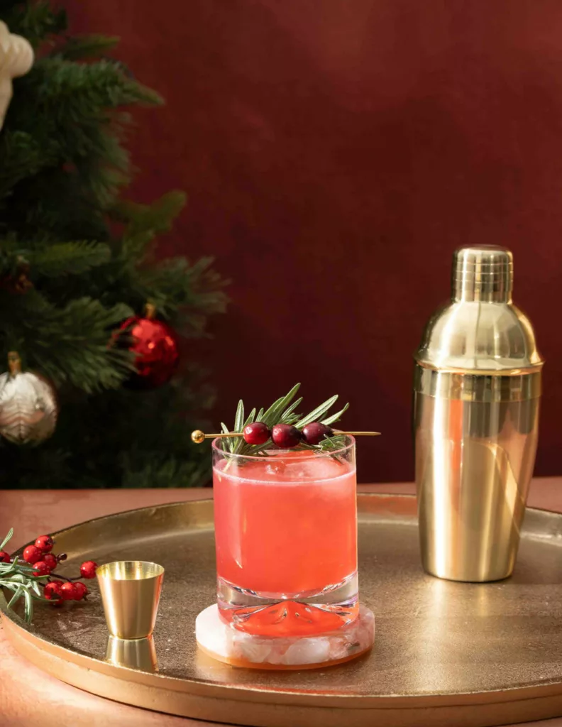 Festive Celosa Winter Spritz cocktail adorned with pomegranate seeds and rosemary, served next to a gold shaker on a golden tray, set against a rich red backdrop with Christmas decorations.