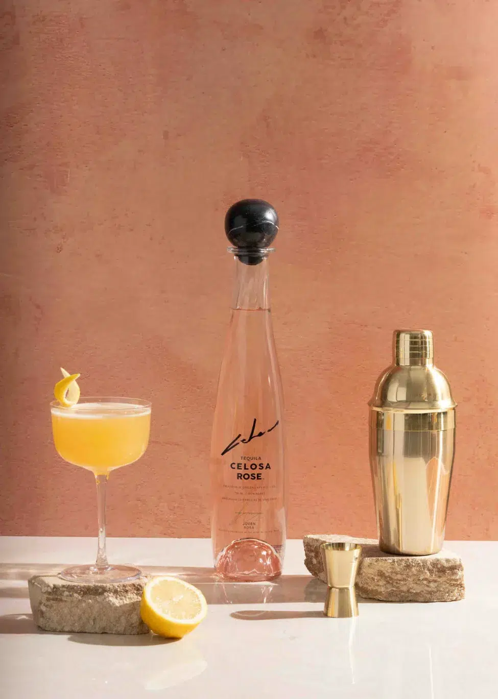 Elegant setup featuring Celosa Rose Tequila bottle alongside a citrusy Celosa Zest cocktail with a lemon twist, a gold cocktail shaker, and jigger on natural stone coasters with a soft terracotta backdrop.