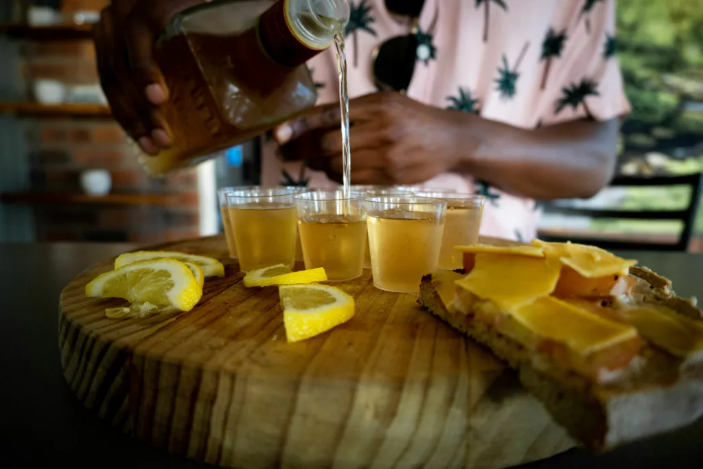 A person is pouring tequila into small glasses arranged on a wooden board that holds slices of lemon.