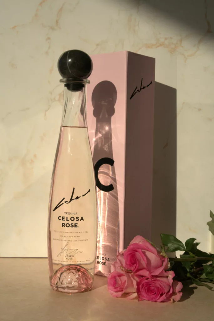 A bottle of Celosa Rosa Tequila with a black cap next to its pink box and roses next to it.