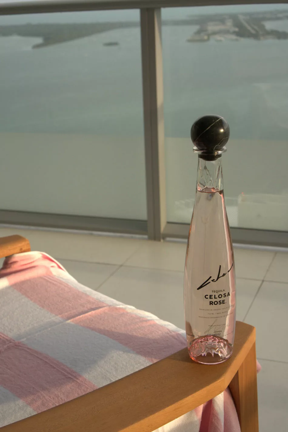 A bottle of Celosa Rosa Tequila, recommended for shots, sits on a chair with an ocean view in the background.