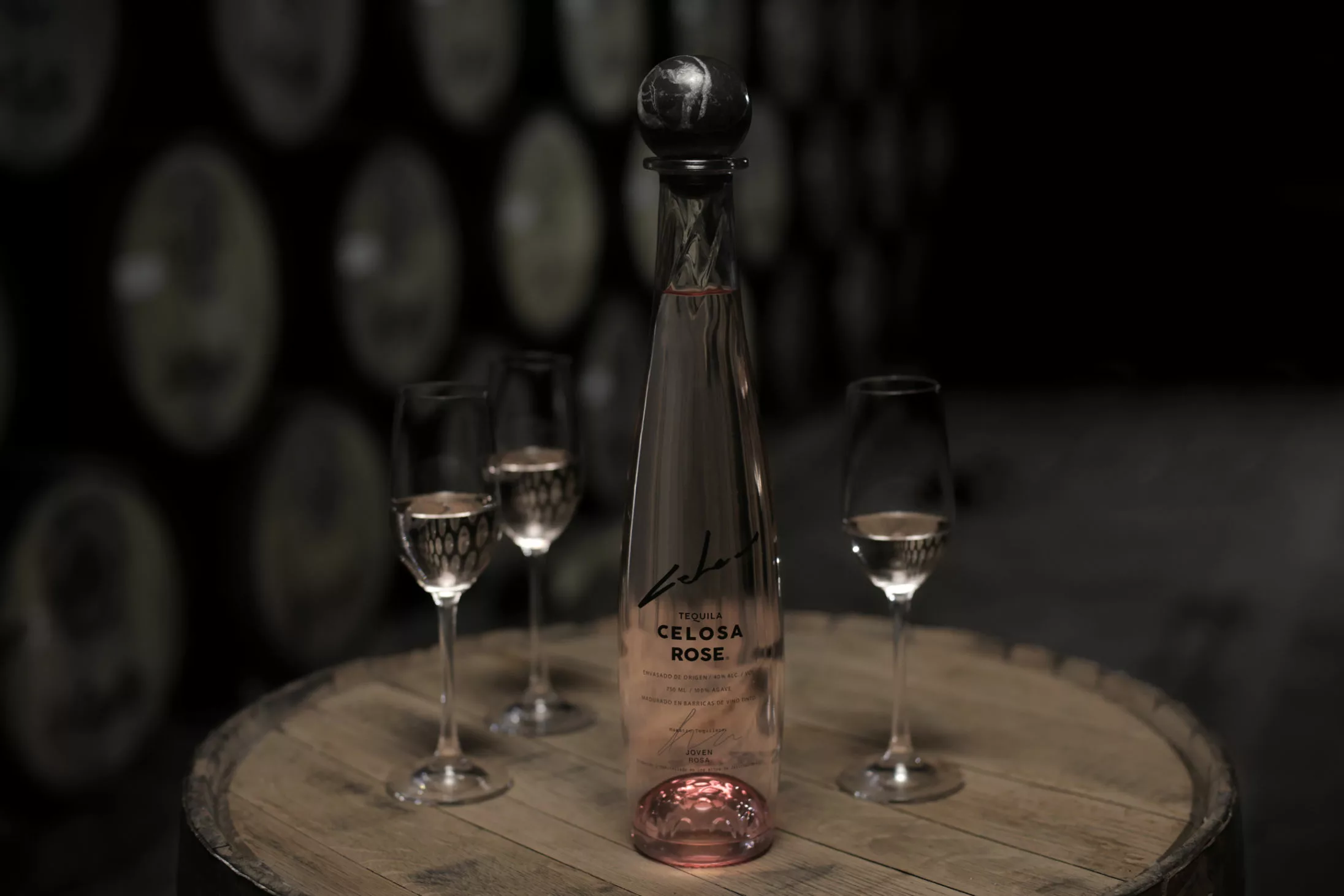 A bottle of Mexican Celosa Rose tequila and three glasses on a wooden barrel, with blurred barrels in the background.