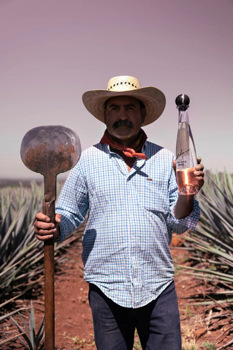 A man in traditional attire holding a hoe and a bottle of tequila stands in an agave field, representing the production of mezcal and tequila.
